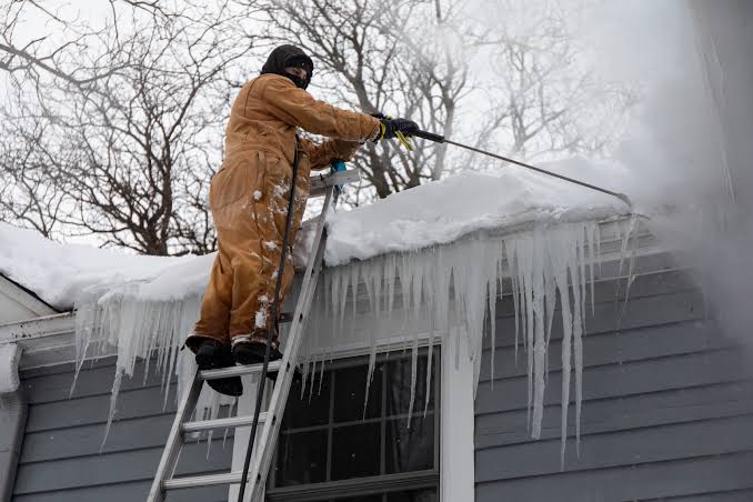Worker in orange coveralls on ladder removing snow and ice dam from roof