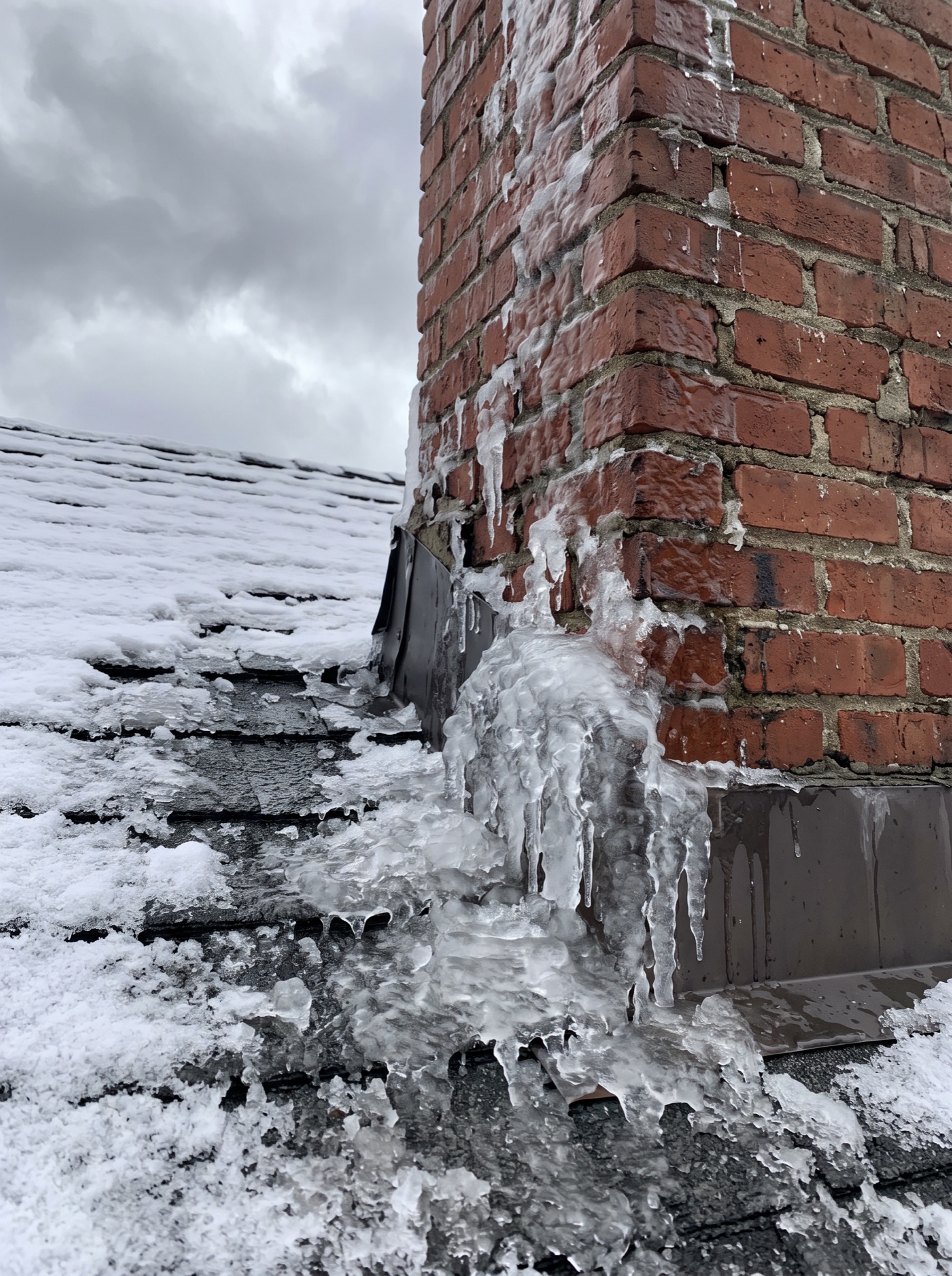 Ice buildup and icicles forming at brick chimney flashing on snowy roof