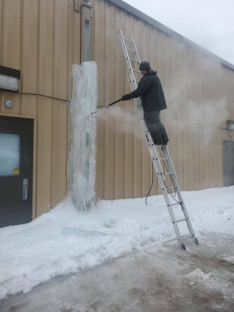 Worker on ladder using steam wand to remove large ice dam from building wall