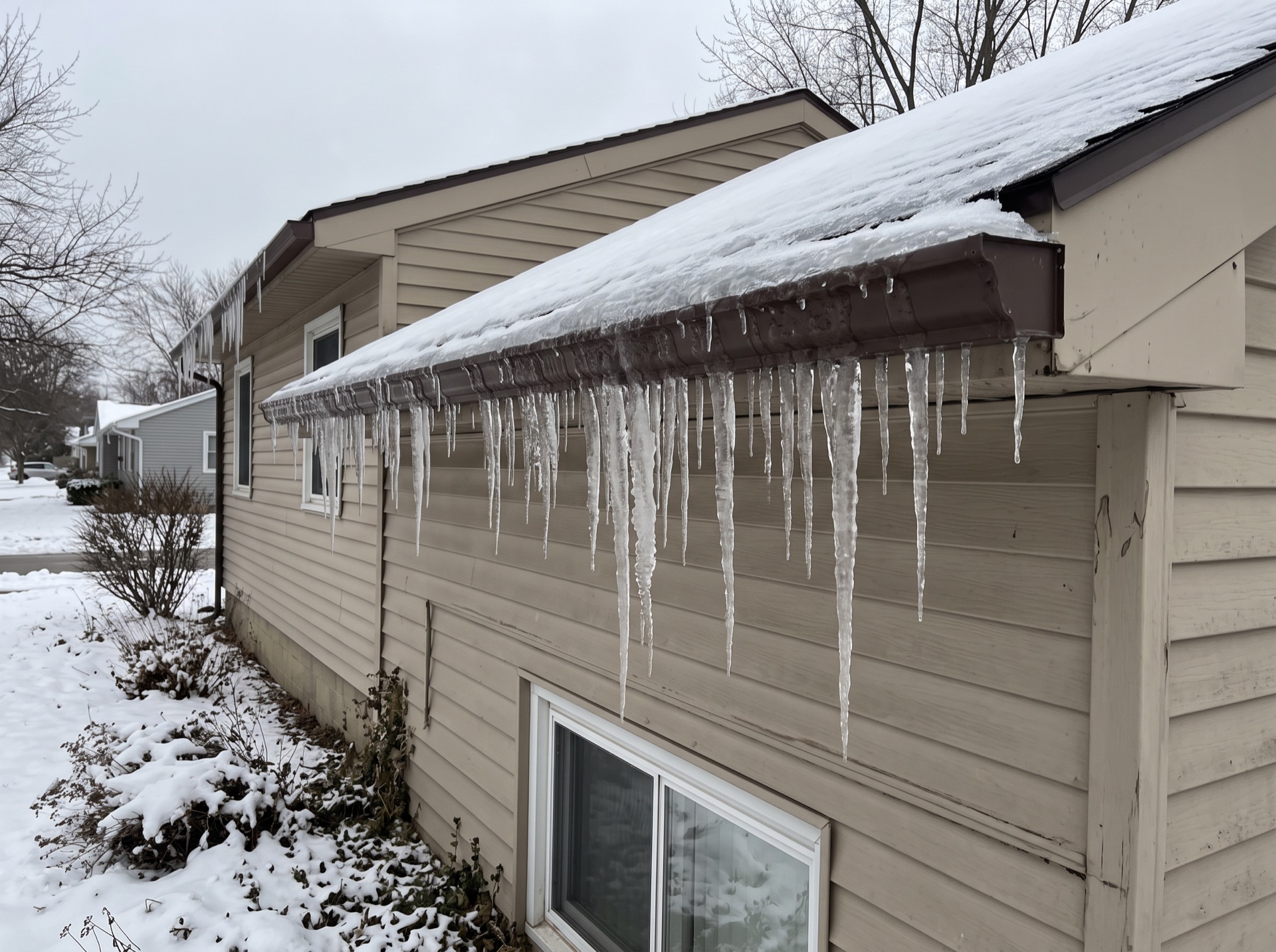 Large icicles and ice dam forming along frozen gutter of beige vinyl siding home