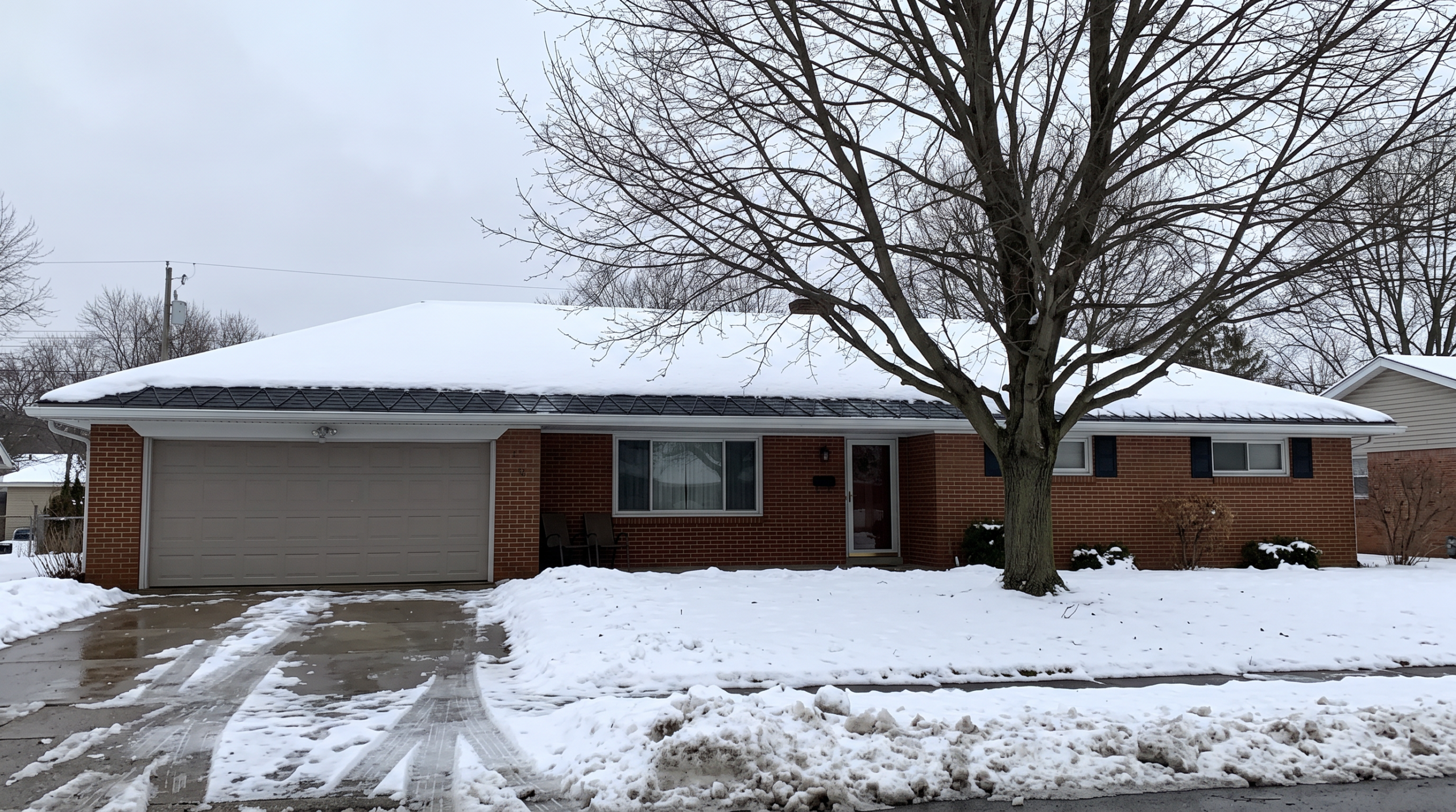 Snow-covered brick ranch home with low-slope roof and bare winter tree