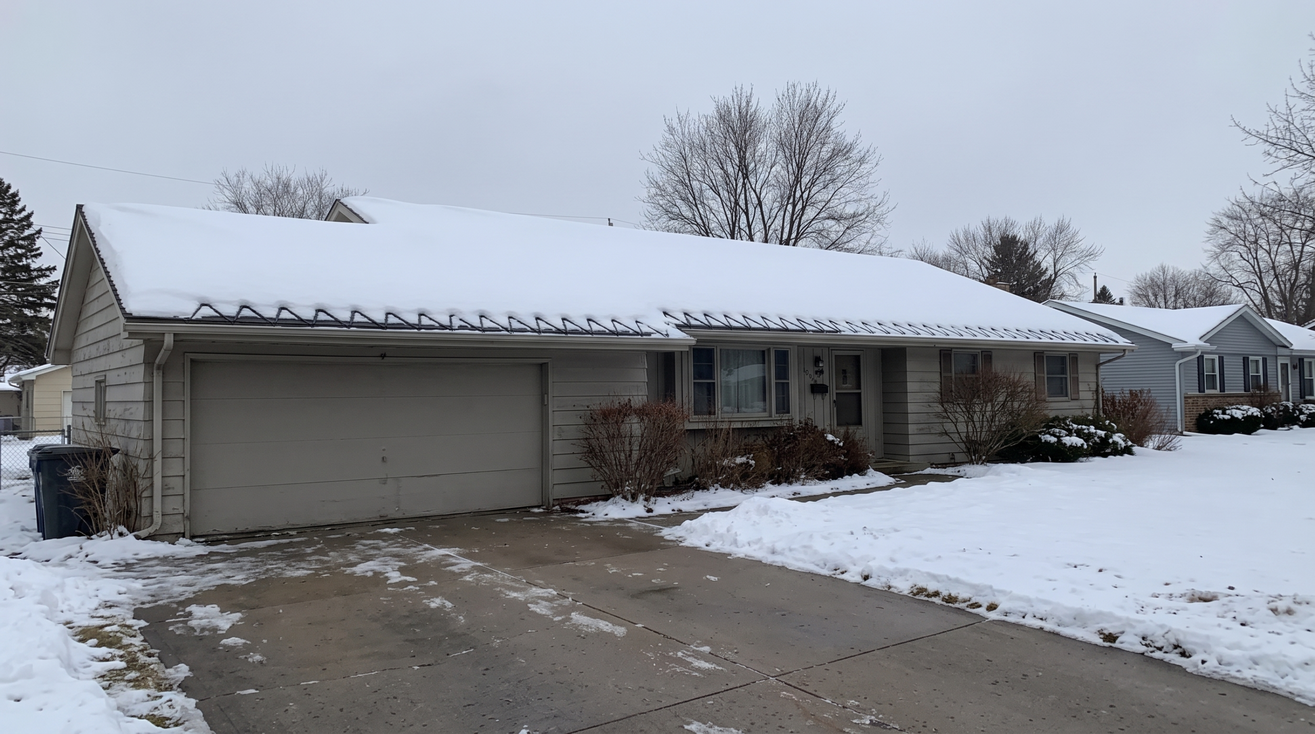Ranch home with ice dam prevention cables along snowy roofline and garage