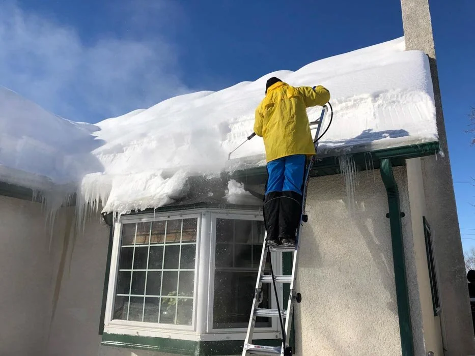 worker in yellow jacket removing snow and ice from rooftop using steam on ladder