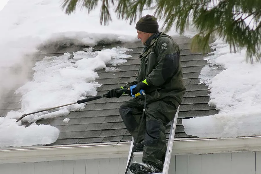 Worker on ladder using steam wand to remove ice dam from snowy roof