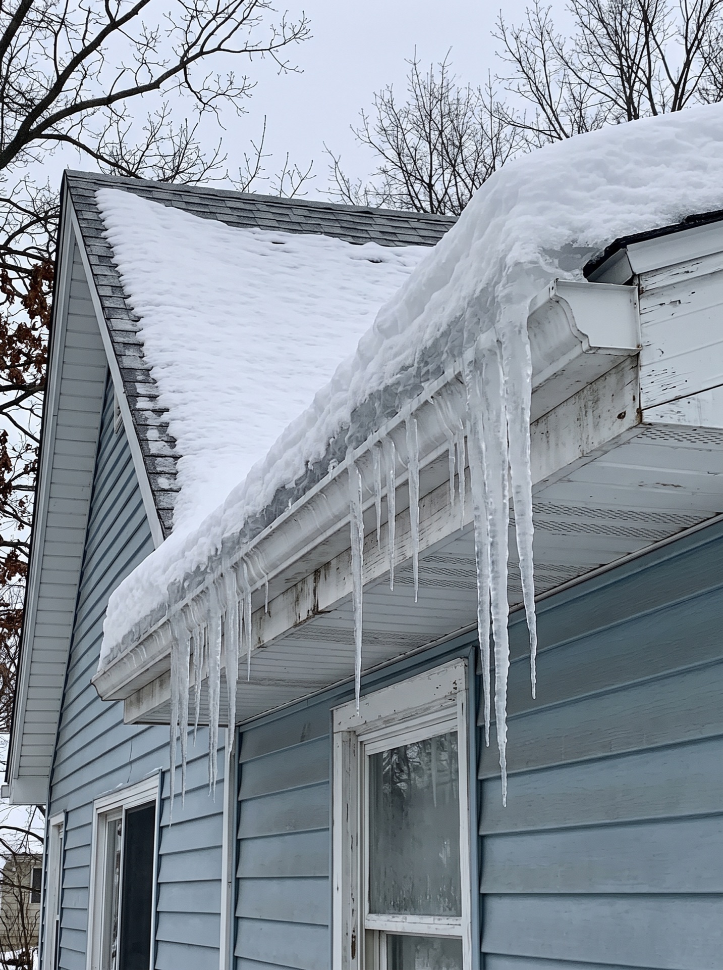 Large ice dam with long icicles hanging from snow-covered roof gutter on blue ranch house