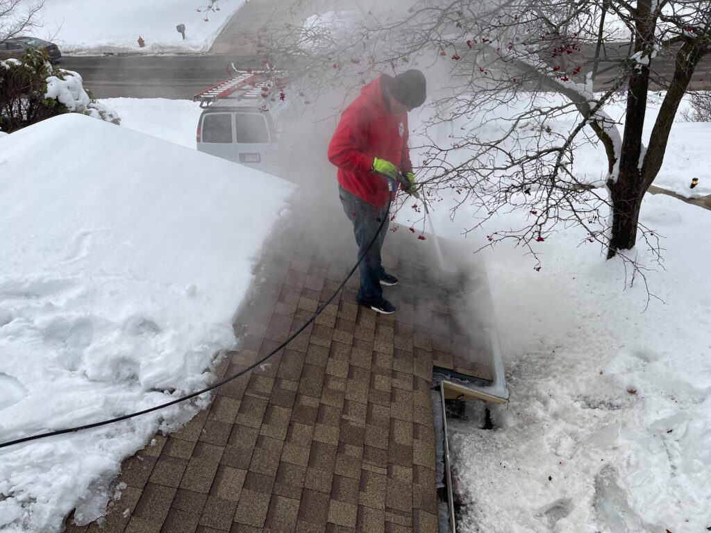 technician using steam lance to remove ice dam from shingle roof eave in winter