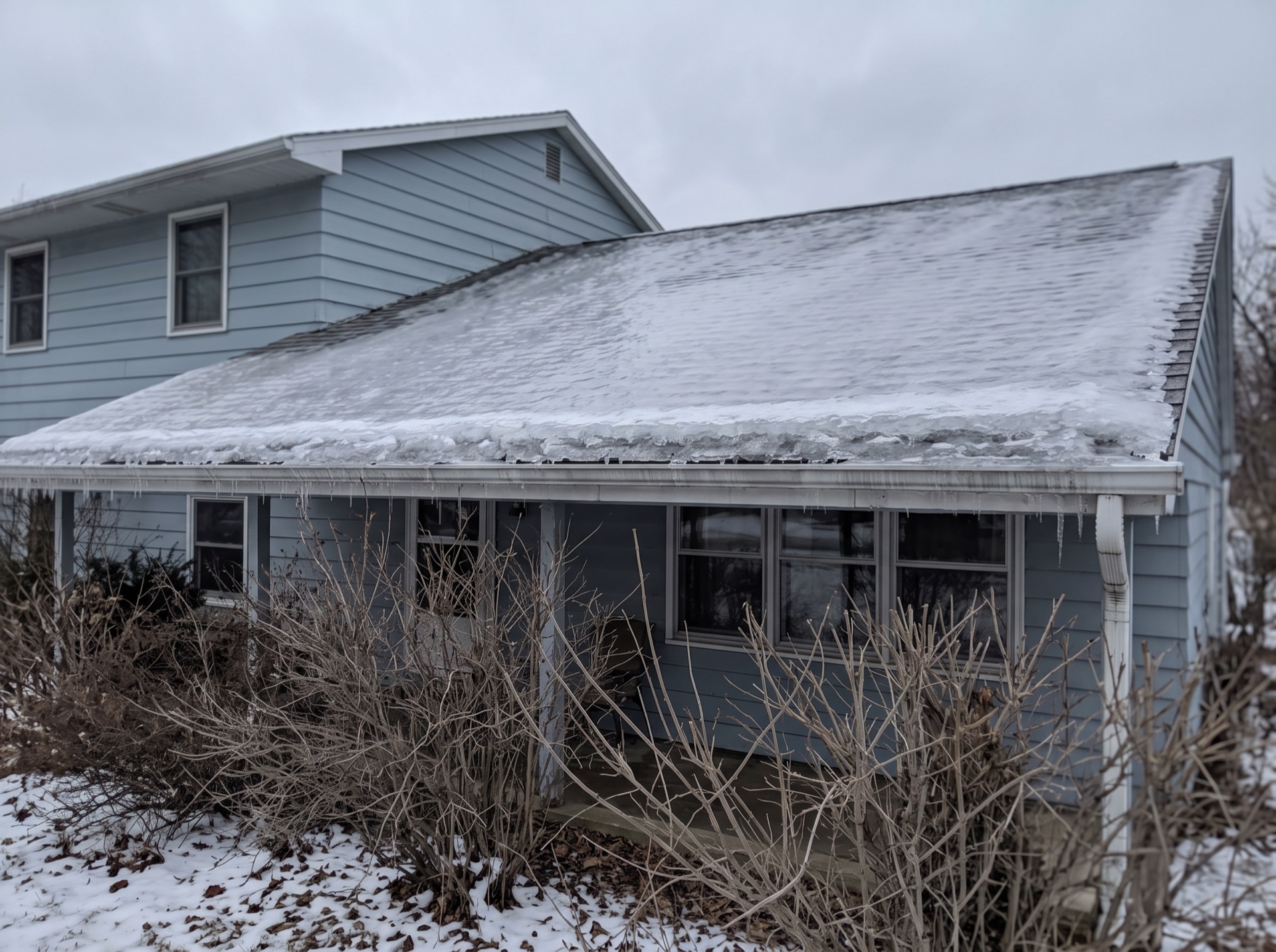 Ice sheet and icicles forming on low slope porch roof of blue house in winter