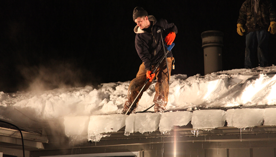 Worker using steam equipment to remove ice dam from snowy roof at night