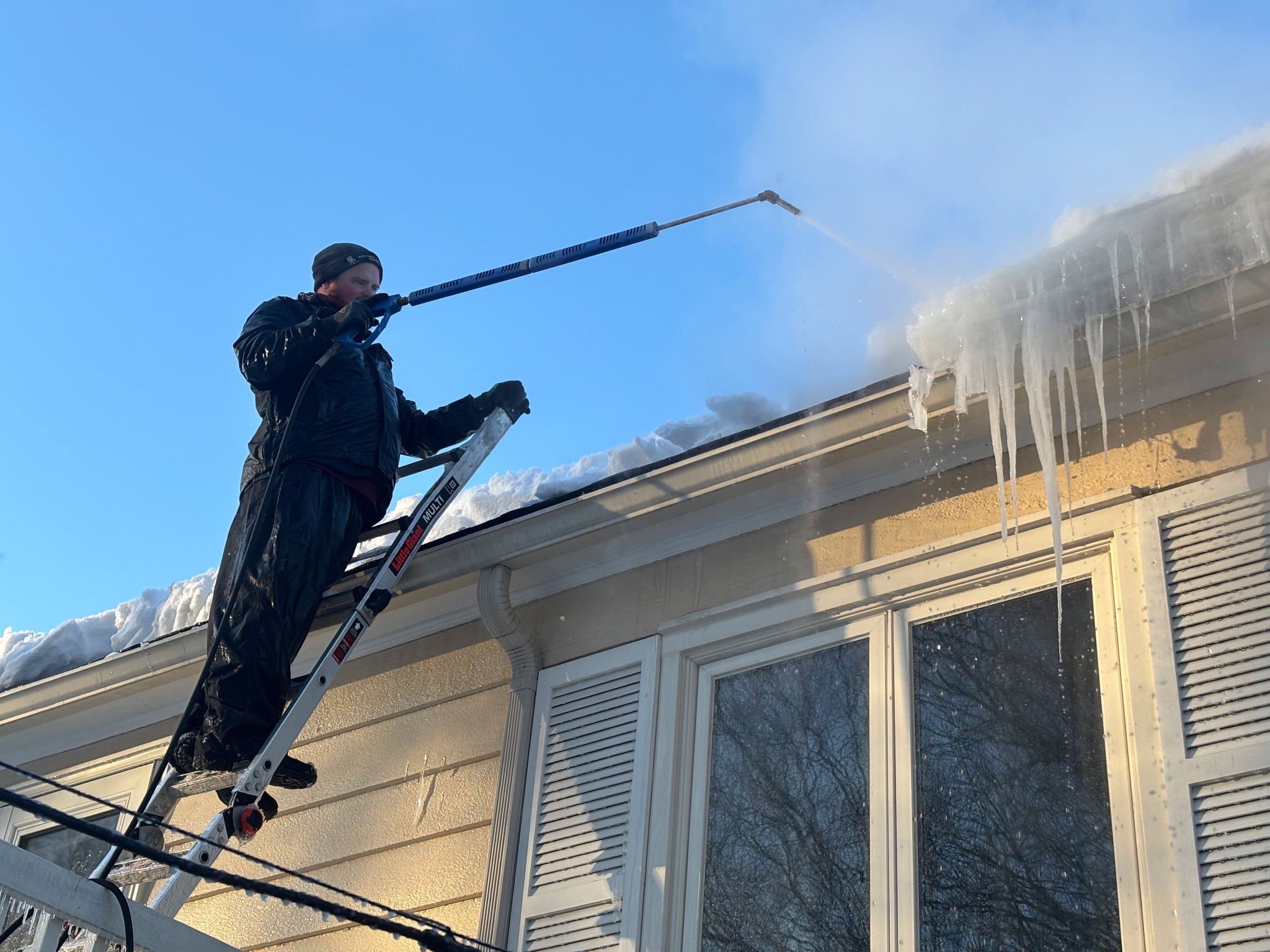 Worker on ladder using steam wand to remove ice dams and icicles from residential roof